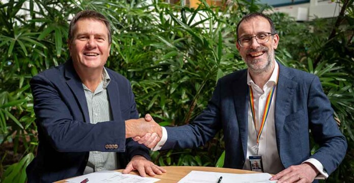 Two male executives in suits shake hands after signing agreement, with greenery in the background